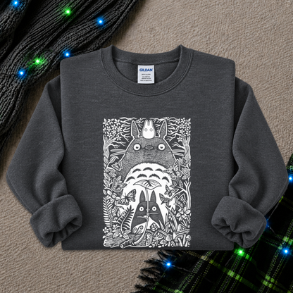 A dark gray crewneck sweatshirt featuring a detailed black and white graphic of Totoro and other My Neighbor Totoro characters, laid flat on a textured beige background with decorative knit items and blue fairy lights.