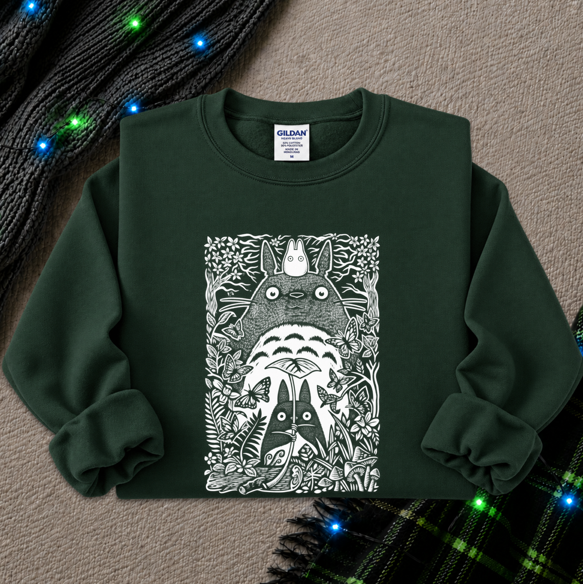 A dark forest green crewneck sweatshirt featuring a large white graphic print of Totoro and other My Neighbor Totoro characters in a detailed, illustrative style. The sweatshirt is laid flat on a textured beige surface, styled with a black knit scarf and a green plaid blanket with glowing blue fairy lights.