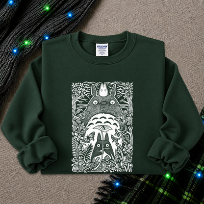 A dark forest green crewneck sweatshirt featuring a large white graphic print of Totoro and other My Neighbor Totoro characters in a detailed, illustrative style. The sweatshirt is laid flat on a textured beige surface, styled with a black knit scarf and a green plaid blanket with glowing blue fairy lights.