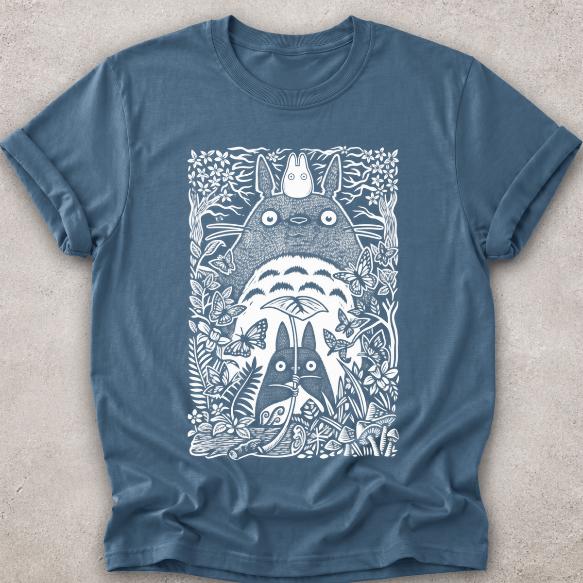 A slate blue crewneck t-shirt featuring a detailed white line art graphic of Totoro and other My Neighbor Totoro characters surrounded by foliage, laid flat on a textured light gray background.