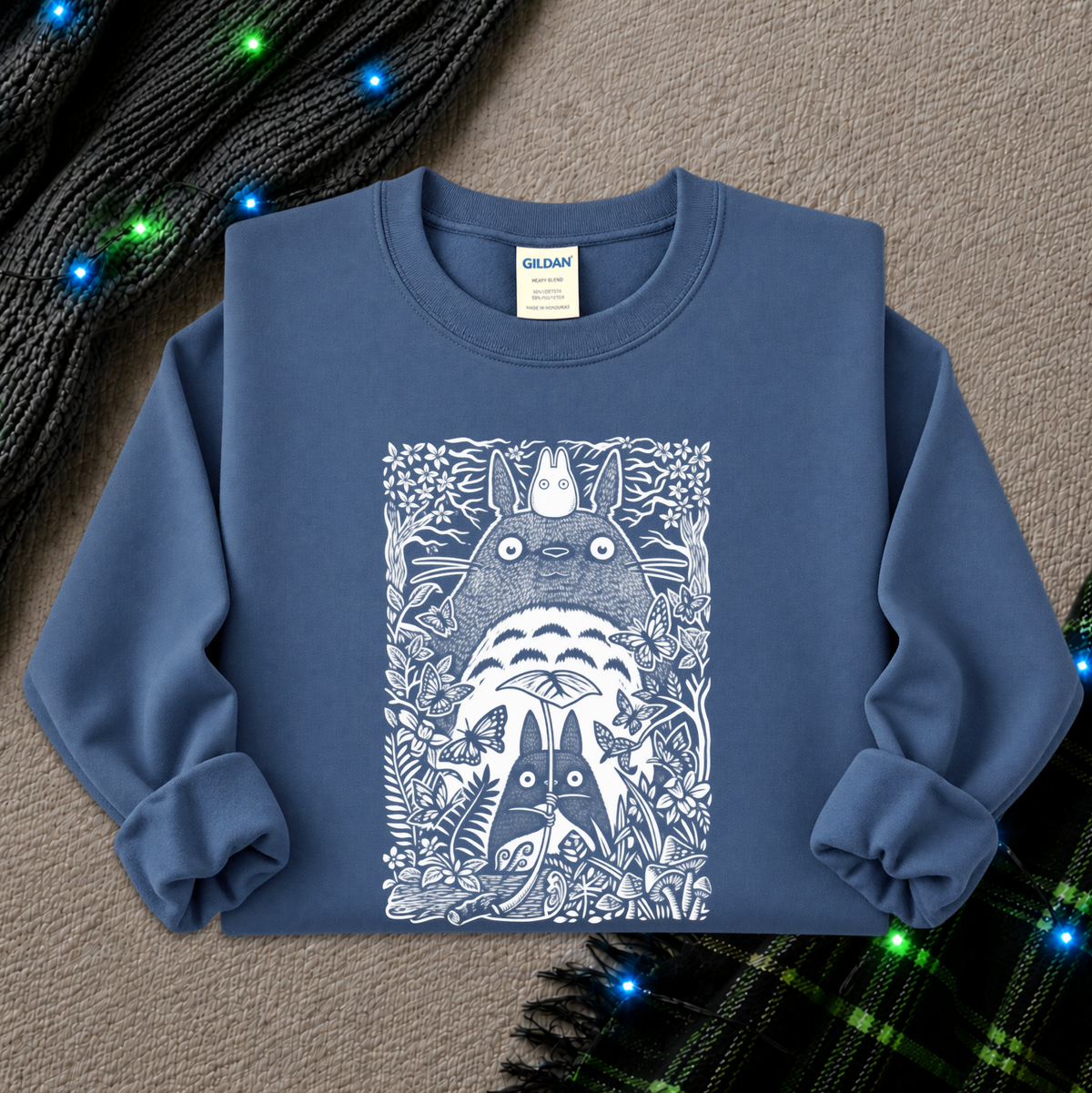 A slate blue crewneck sweatshirt featuring a white line art graphic of Totoro and other My Neighbor Totoro characters, styled flat on a textured beige background with holiday lights and a plaid scarf.