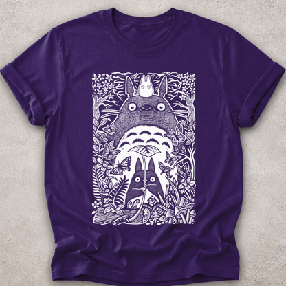 A purple short-sleeve crewneck t-shirt featuring a white graphic print of Totoro and other My Neighbor Totoro characters surrounded by detailed foliage, laid flat on a textured light gray background.