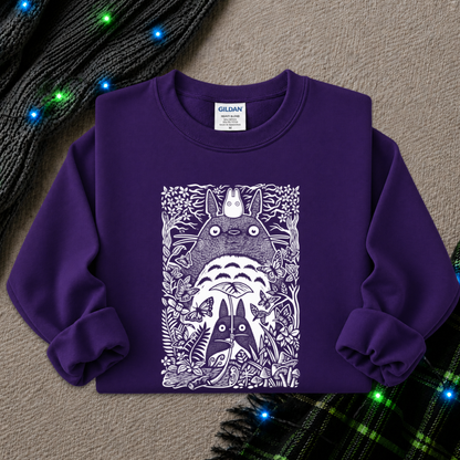 A purple long-sleeve crewneck sweatshirt featuring a white graphic print of Totoro and other My Neighbor Totoro characters, laid flat on a textured beige background with holiday lights and knit fabric accents.