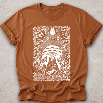 A rust-brown short-sleeve t-shirt with a white screen-printed graphic of Totoro and other My Neighbor Totoro characters surrounded by foliage, laid flat on a textured gray background.