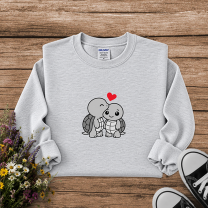 A light gray crewneck sweatshirt with a cute cartoon turtle graphic on the front, styled in a flat lay on a wooden background with flowers and black sneakers.
