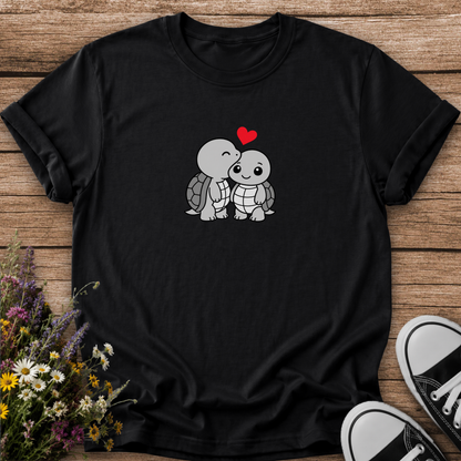 A black short-sleeve t-shirt laid flat on a rustic wooden surface, featuring a cute cartoon graphic of two gray turtles hugging with a small red heart above them. The shirt is styled with a bouquet of wildflowers on the left and black-and-white sneakers on the right.