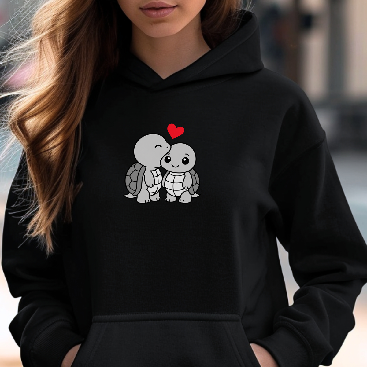 A woman wearing a black pullover hoodie with a cute cartoon graphic of two kissing sea turtles under a small red heart on the chest.