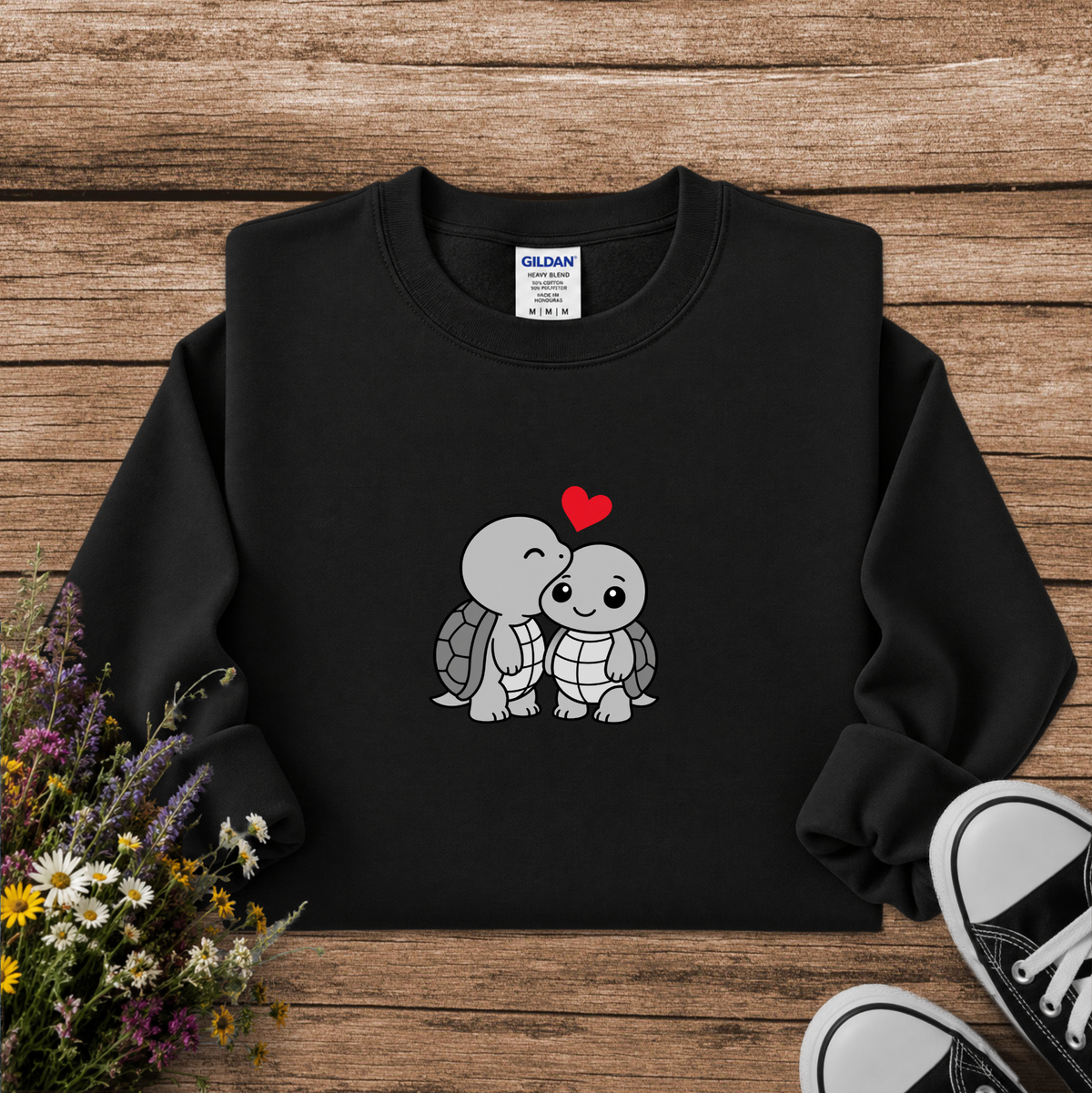A black crewneck sweatshirt featuring a cute cartoon graphic of two kissing sea turtles with a small red heart above them. The sweatshirt is displayed in a flat-lay arrangement on a rustic wooden background, styled with a bouquet of wildflowers and the corner of a black canvas sneaker.