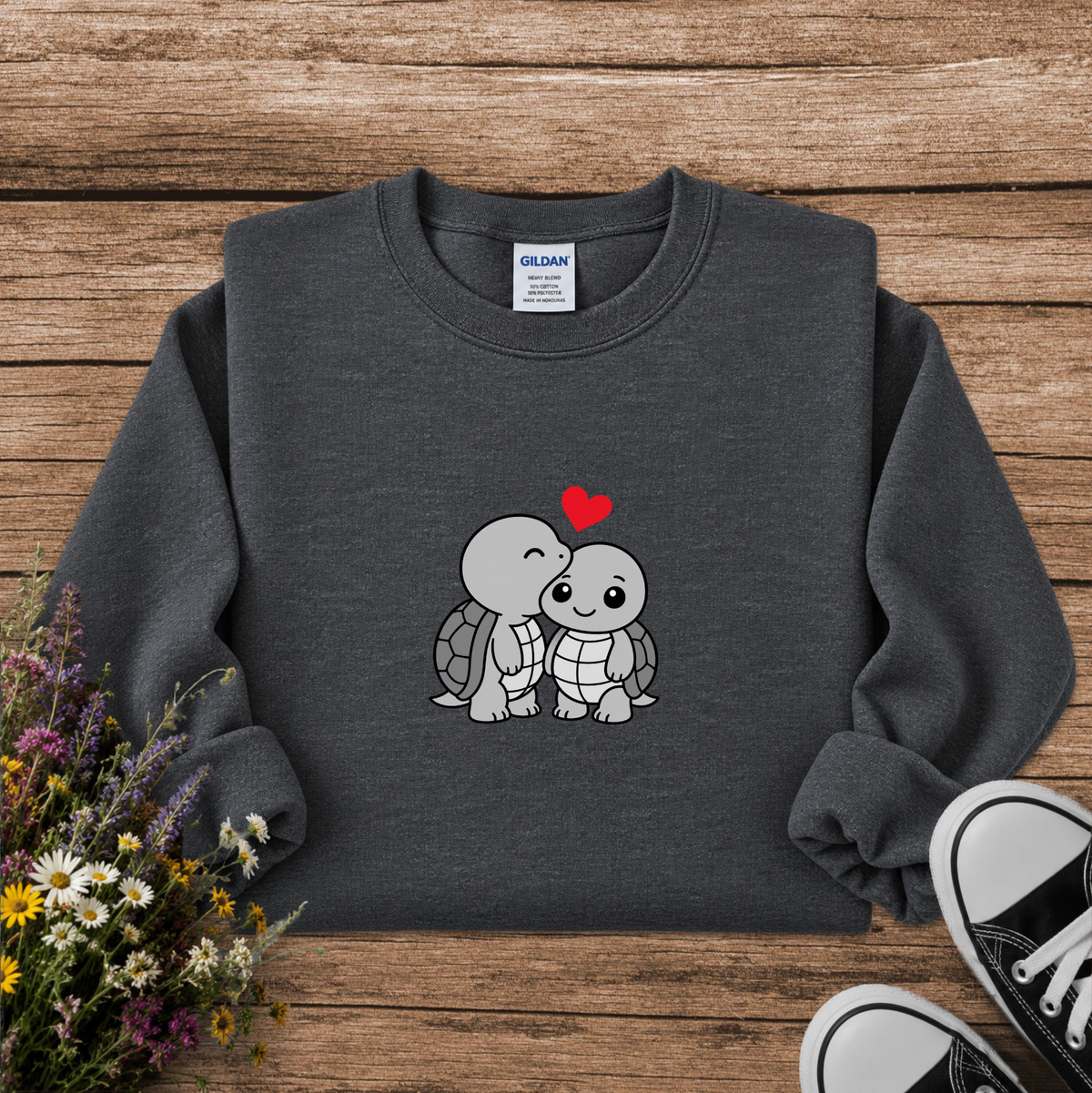 A dark gray crewneck sweatshirt featuring a cute cartoon graphic of two hugging turtles with a small red heart above them. The sweatshirt is laid flat on a rustic wooden background, styled with a small bouquet of wildflowers on the left and the corner of black canvas sneakers on the bottom right.