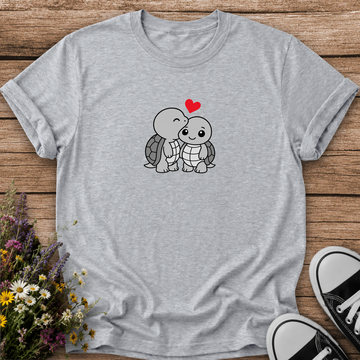 A light heather gray short-sleeve t-shirt with a cute cartoon turtle graphic on the chest, styled in a flat-lay on a wooden surface with flowers and black-and-white sneakers.