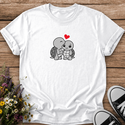 A white short-sleeve crewneck t-shirt laid flat on a rustic wooden surface, featuring a cute cartoon graphic of two kissing turtles with a small red heart above them. The shirt is styled with wildflowers on the left and black-and-white sneakers on the right.