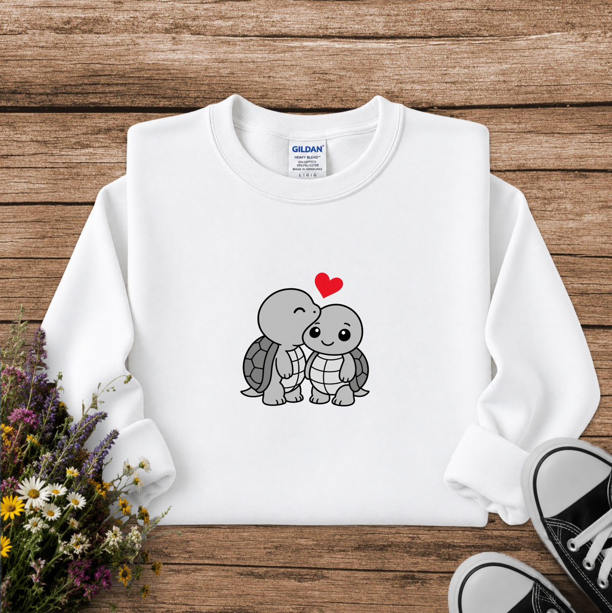 A white long-sleeve crewneck sweatshirt with a cute cartoon turtle graphic on the chest, styled in a flat lay on a rustic wooden background with wildflowers and black sneakers.