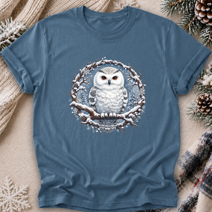 A slate blue crewneck t-shirt featuring a detailed graphic of a snowy owl perched on a snow-dusted branch inside a circular wreath. The t-shirt is styled in a cozy flat-lay setting with a beige knit sweater, pinecones, and a plaid scarf.
