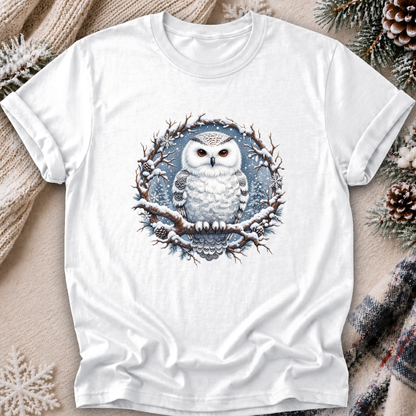 A white short-sleeve crewneck t-shirt featuring a detailed graphic of a snowy owl perched on a snow-dusted branch. The shirt is presented in a flat-lay arrangement with cozy holiday props including a knit sweater, pinecones, and a plaid scarf on a neutral beige background.