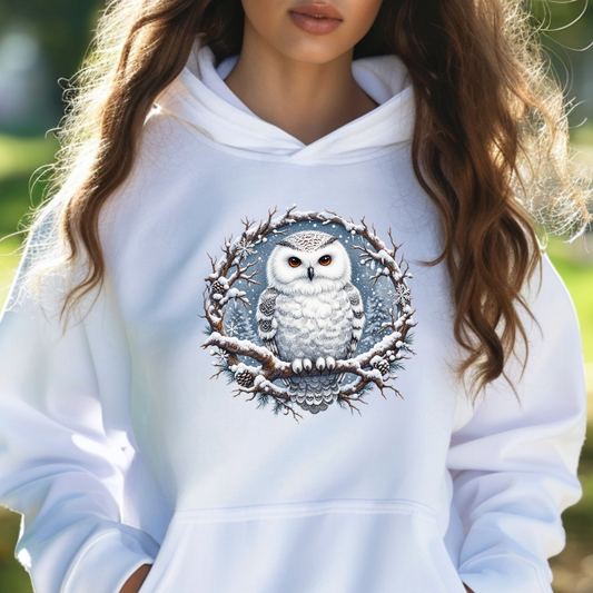 A woman wearing a white pullover hoodie with a detailed graphic of a snowy owl framed by a snow-covered tree branch wreath on the chest.