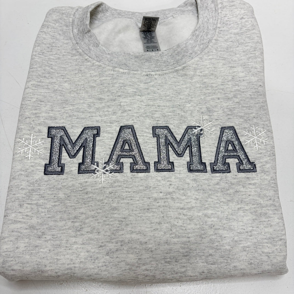 A folded light heather gray sweatshirt featuring the word 'MAMA' in dark gray varsity-style block letters with white glitter-like fill and small white snowflakes on either side, shown on a plain white background.