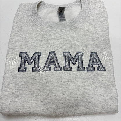 A folded light heather gray sweatshirt featuring the word 'MAMA' in dark gray varsity-style block letters with white glitter-like fill and small white snowflakes on either side, shown on a plain white background.