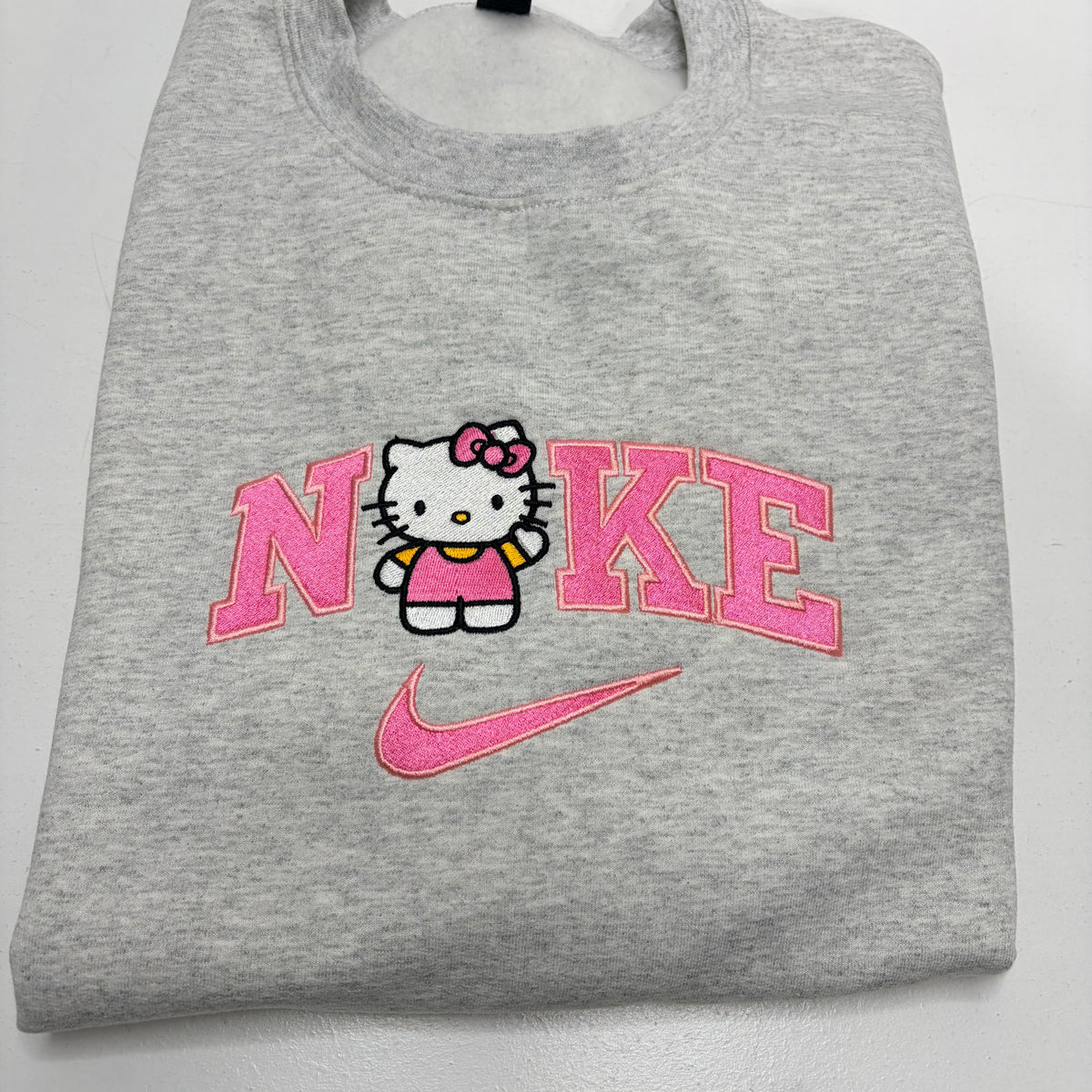 A folded heather gray sweatshirt featuring a pink and white embroidered design on the front with the word 'NIKE' stylized around Hello Kitty, and a pink Nike swoosh below.