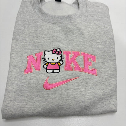 A folded heather gray sweatshirt featuring a pink and white embroidered design on the front with the word 'NIKE' stylized around Hello Kitty, and a pink Nike swoosh below.