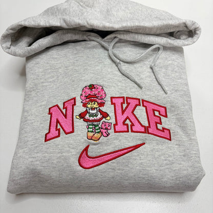 A folded light gray heather hoodie with a pink and red embroidered design on the front. The design features the word 'NIKE' in large pink letters with a cartoon strawberry shortcake character in the center and a pink Nike swoosh below.