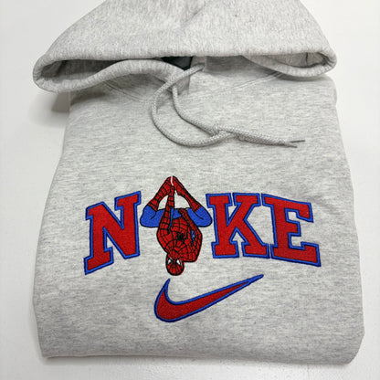 A folded light gray heather hoodie featuring a Spider-Man graphic and the word 'NOKE' with a red and blue Nike swoosh underneath.