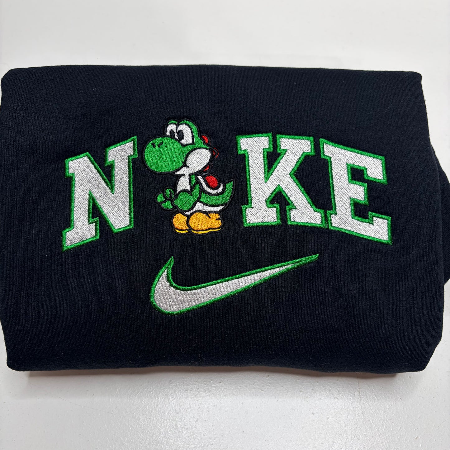 A folded black sweatshirt featuring a custom embroidered design with the word 'N'KE' in white varsity letters outlined in green, a green Yoshi character in the center, and a green Nike swoosh below.