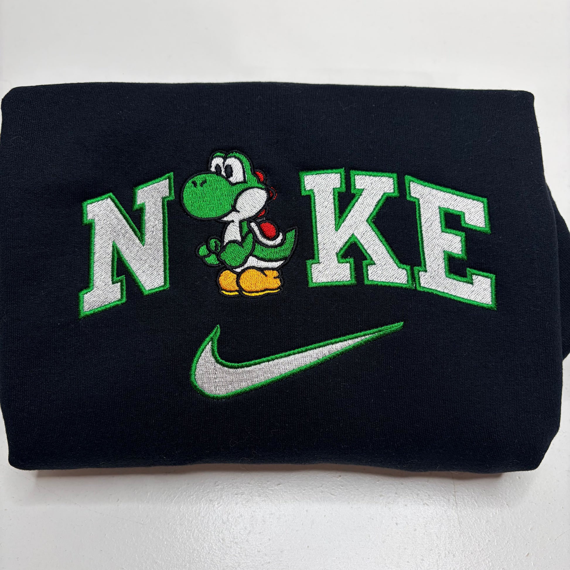 A folded black sweatshirt featuring a custom embroidered design with the word 'N'KE' in white varsity letters outlined in green, a green Yoshi character in the center, and a green Nike swoosh below.