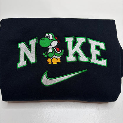 A folded black sweatshirt featuring a custom embroidered design with the word 'N'KE' in white varsity letters outlined in green, a green Yoshi character in the center, and a green Nike swoosh below.