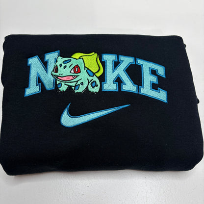 A folded black sweatshirt featuring a light blue embroidered 'NIKE' logo, a Bulbasaur character graphic, and a matching Nike swoosh on the front.
