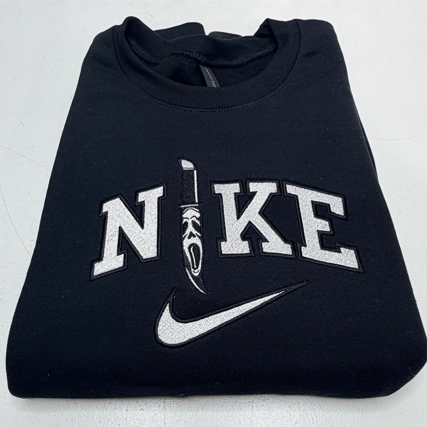 A folded black Nike sweatshirt featuring a large white embroidered 'NIKE' logo with a scream ghost knife graphic and a white swoosh on the front.