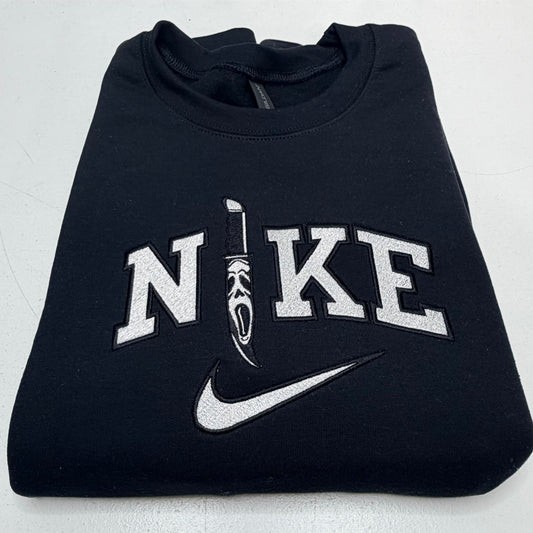 A folded black Nike sweatshirt featuring a large white embroidered 'NIKE' logo with a scream ghost knife graphic and a white swoosh on the front.