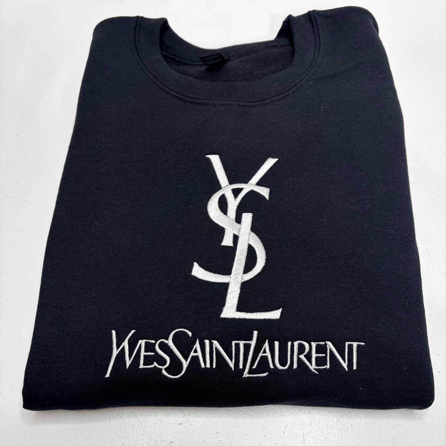 A folded black crewneck t-shirt featuring a large white embroidered Yves Saint Laurent YSL logo and 'Yves Saint Laurent' text on the front, set against a plain white background.