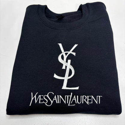 A folded black crewneck t-shirt featuring a large white embroidered Yves Saint Laurent YSL logo and 'Yves Saint Laurent' text on the front, set against a plain white background.
