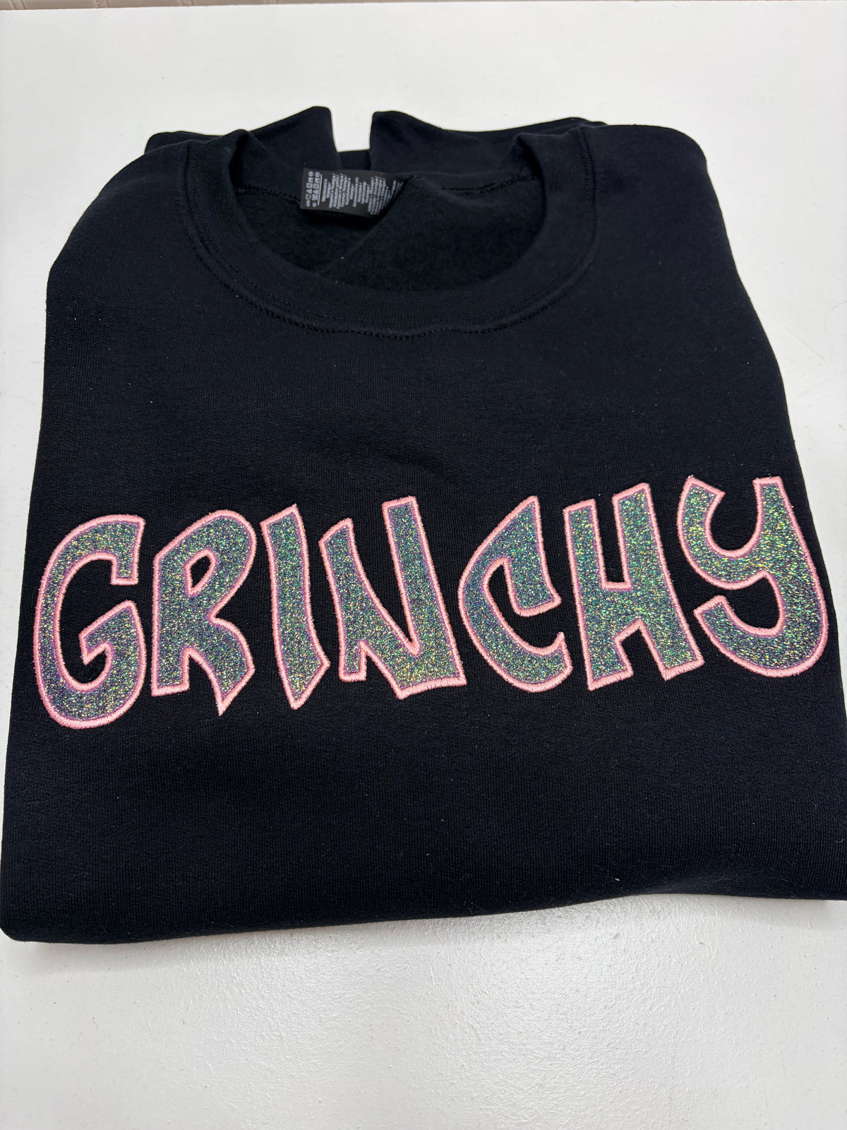 A folded black crewneck sweatshirt featuring the word 'GRINCHY' in large, glittery pink-outlined letters with a green glitter fill, displayed on a light gray background.