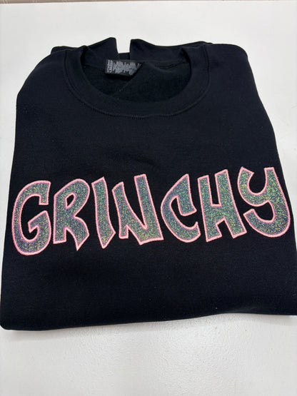 A folded black crewneck sweatshirt featuring the word 'GRINCHY' in large, glittery pink-outlined letters with a green glitter fill, displayed on a light gray background.
