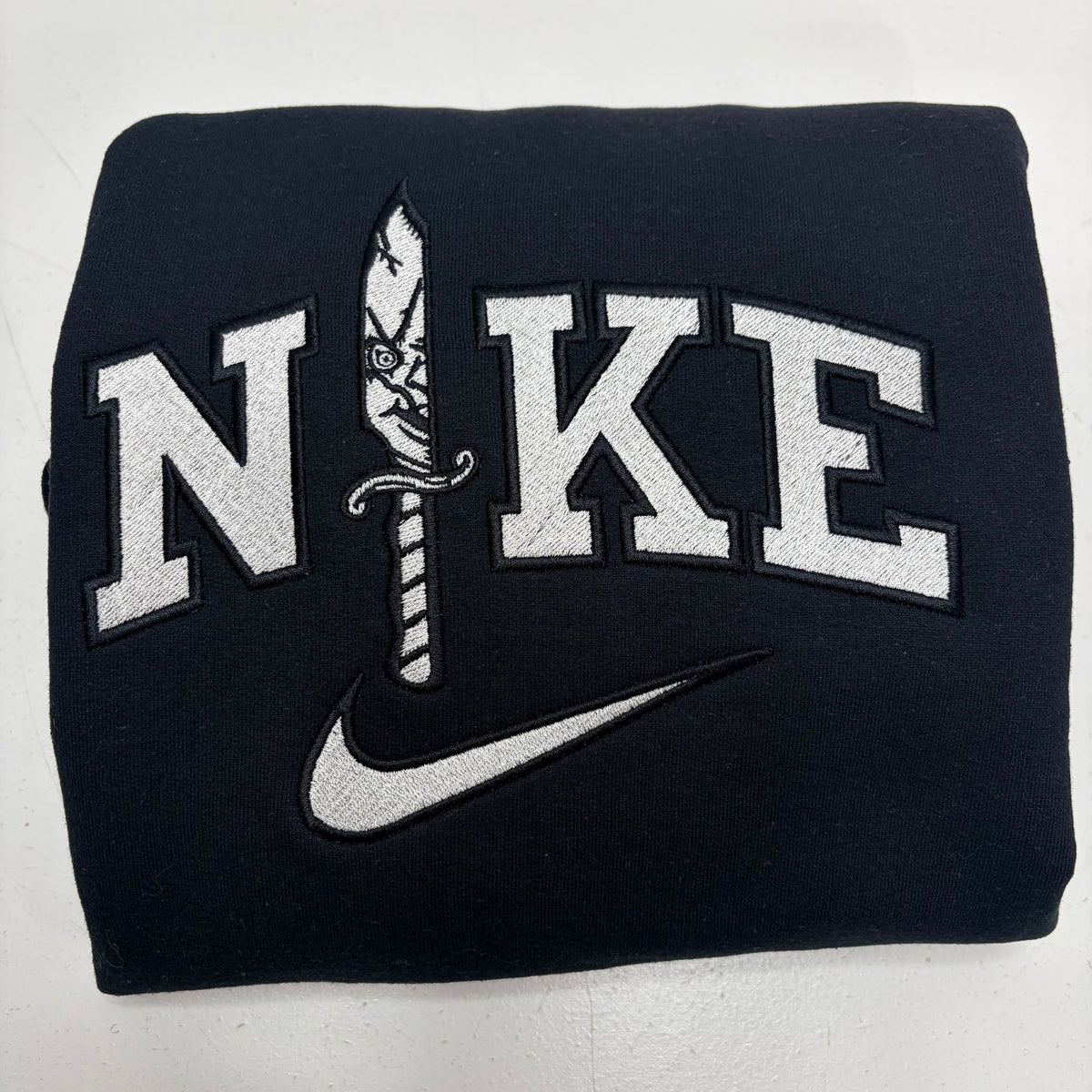 A folded black sweatshirt featuring a large white embroidered 'NIKE' logo with a dagger motif and a white swoosh underneath, shown on a light gray background.