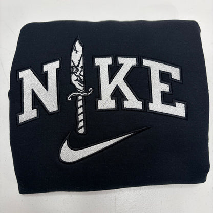 A folded black sweatshirt featuring a large white embroidered 'NIKE' logo with a dagger motif and a white swoosh underneath, shown on a light gray background.