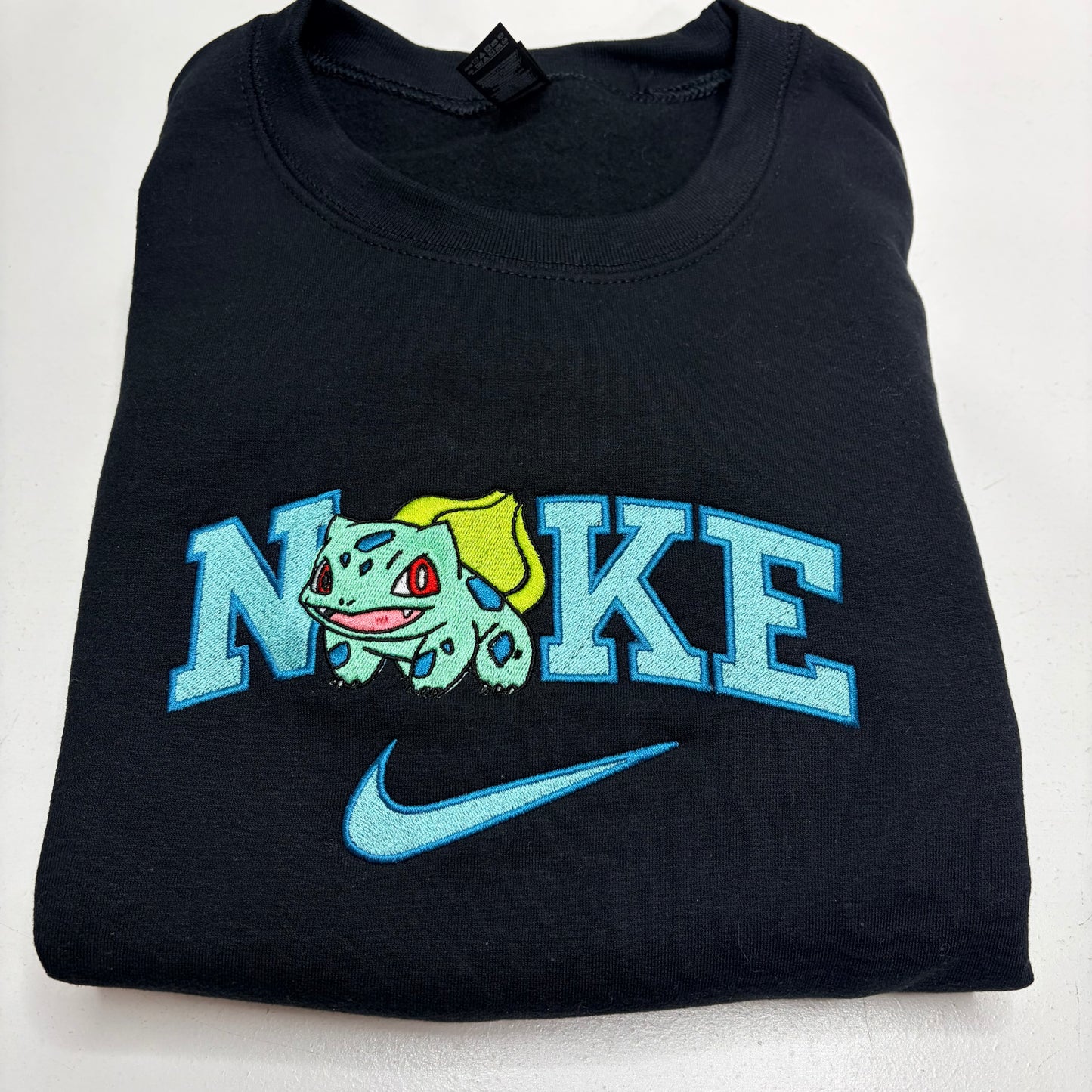 A folded black t-shirt featuring a custom embroidered design. The design spells 'NOKE' with the letter 'O' replaced by a green and yellow Bulbasaur character, and a light blue Nike swoosh is embroidered below.