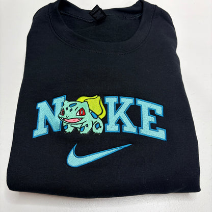 A folded black t-shirt featuring a custom embroidered design. The design spells 'NOKE' with the letter 'O' replaced by a green and yellow Bulbasaur character, and a light blue Nike swoosh is embroidered below.