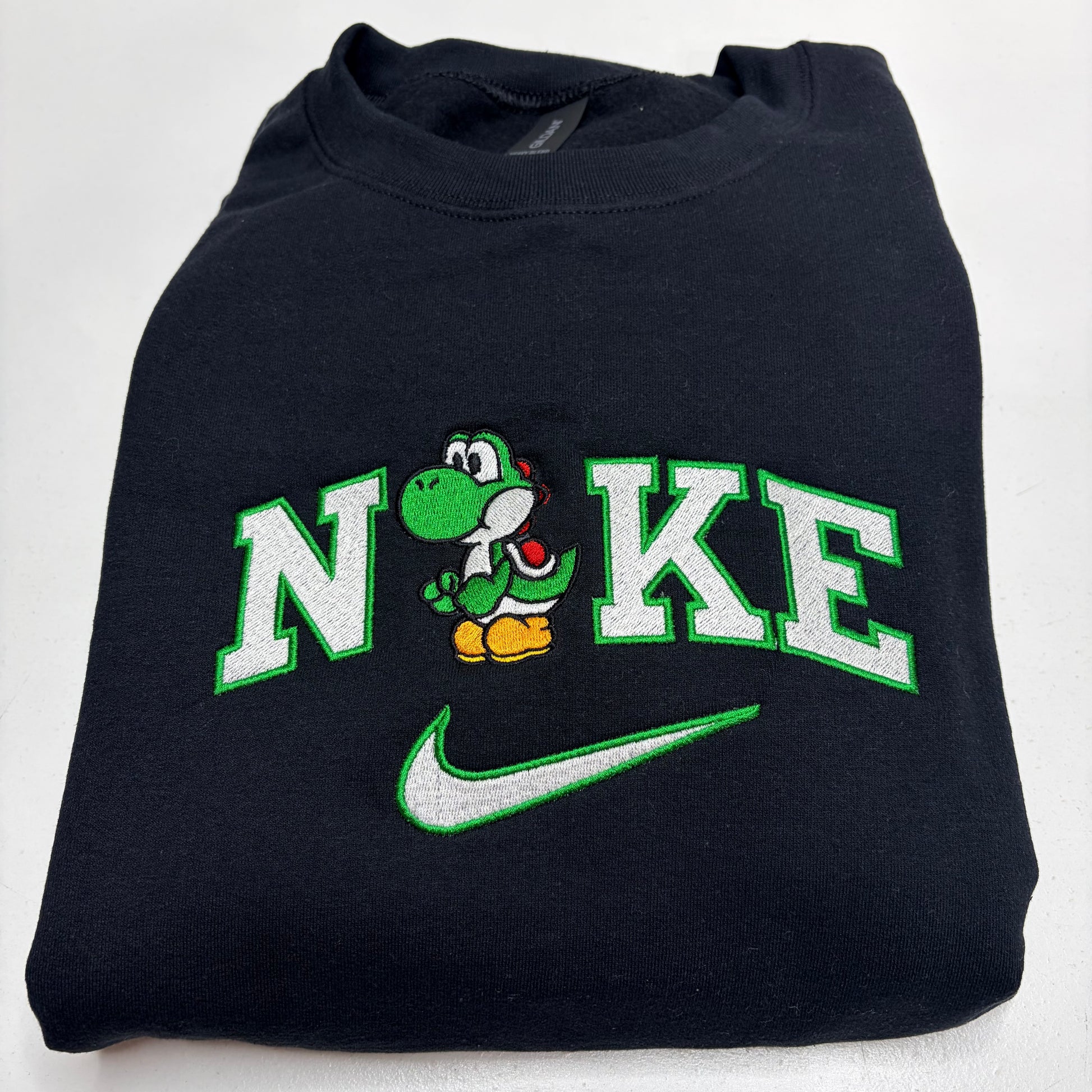 A folded black sweatshirt featuring a custom embroidered design that reads 'N' and 'KE' with a green cartoon Yoshi character in the middle, and a green and white Nike swoosh underneath.