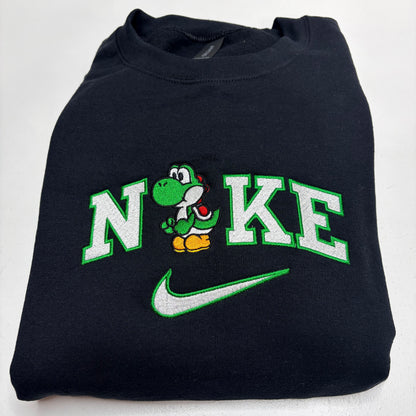 A folded black sweatshirt featuring a custom embroidered design that reads 'N' and 'KE' with a green cartoon Yoshi character in the middle, and a green and white Nike swoosh underneath.