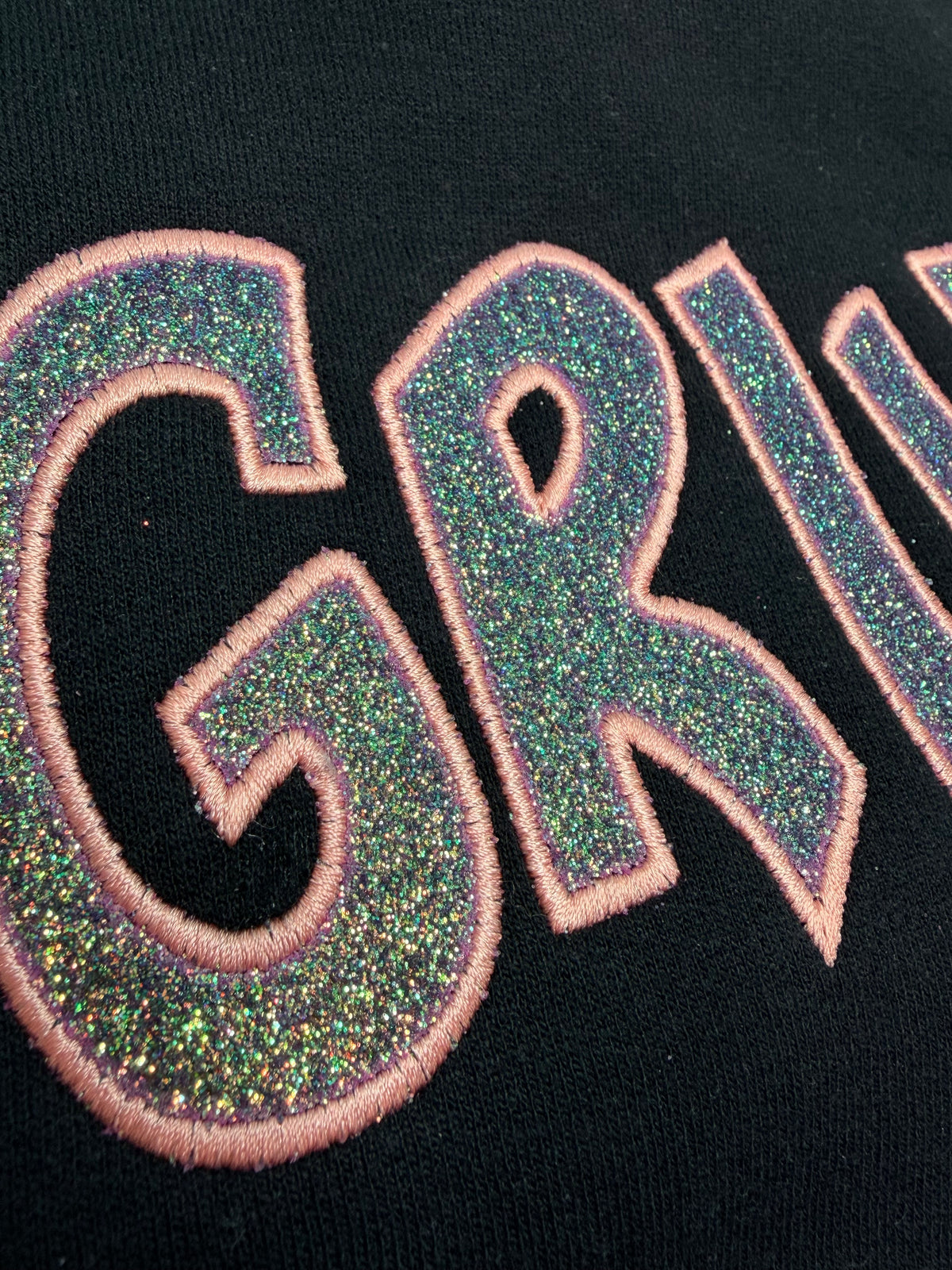Close-up of a black garment featuring large glittery green and blue embroidered letters outlined in pink.