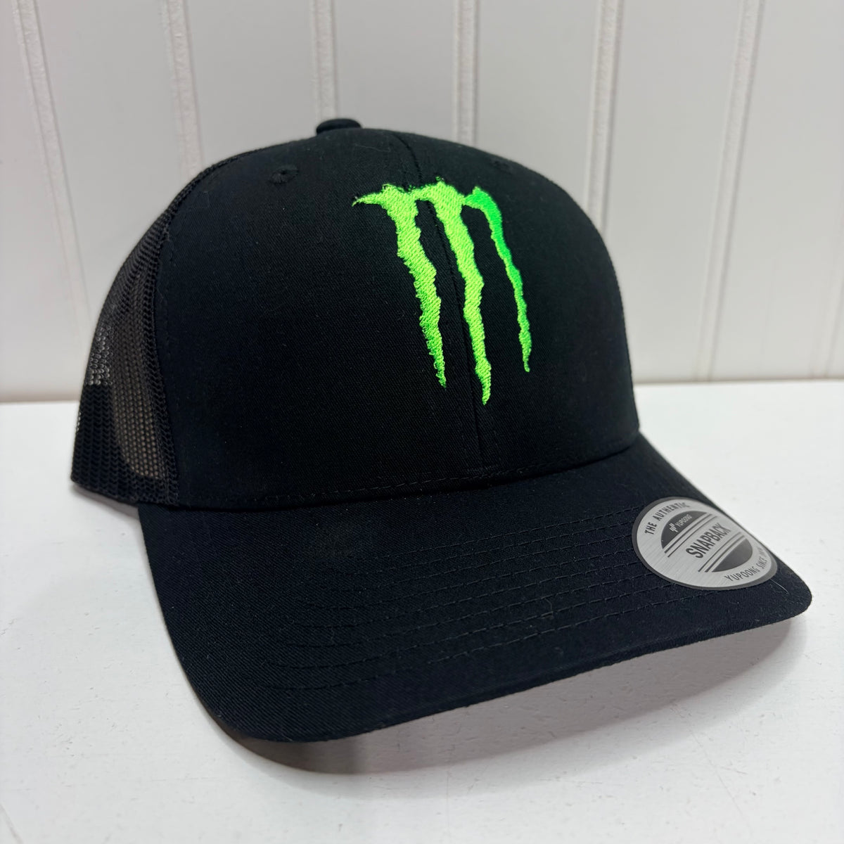 A black trucker-style baseball cap with a bright neon green embroidered Monster Energy claw logo on the front. The cap features black mesh back panels and a curved brim with a silver size sticker on the side, set against a white paneled background.