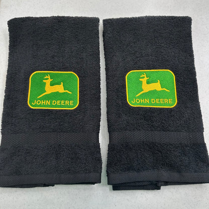 Two black towels with green and yellow John Deere logos on the front, displayed side-by-side on a light gray surface.