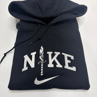 A folded black Nike hoodie featuring a custom embroidered design on the front. The design spells 'NIKE' in white varsity-style block letters, with the letter 'I' replaced by a detailed white and black dagger motif, and a white Nike swoosh embroidered underneath.