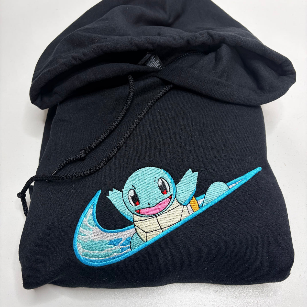 A folded black hoodie featuring an embroidered design of Squirtle riding a blue wave on a Nike swoosh, set against a plain white background.
