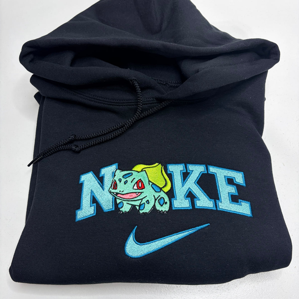A folded black Nike hoodie featuring a custom embroidered design with the word 'NIKE' stylized with Bulbasaur, and a light blue Nike swoosh below.