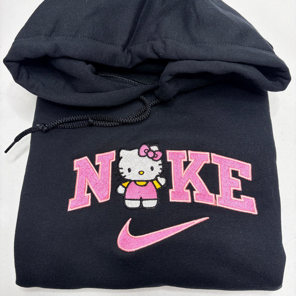 A folded black hoodie featuring a pink and white Hello Kitty graphic with the word 'NIKE' and a pink swoosh on the front.