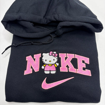 A folded black hoodie featuring a pink and white Hello Kitty graphic with the word 'NIKE' and a pink swoosh on the front.