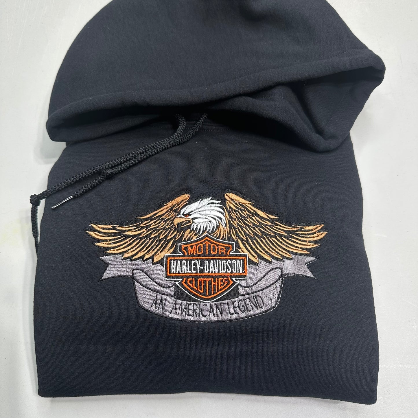 A black pullover hoodie featuring a large embroidered Harley-Davidson winged logo patch on the front, set against a plain light background.